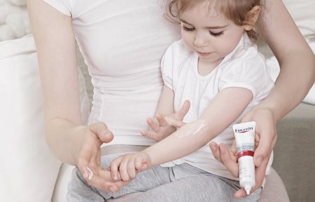 Une petite fille, assise sur les genoux de sa mère, se met de la crème calmante intensive Eucerin AtopiControl dans le pli du coude.